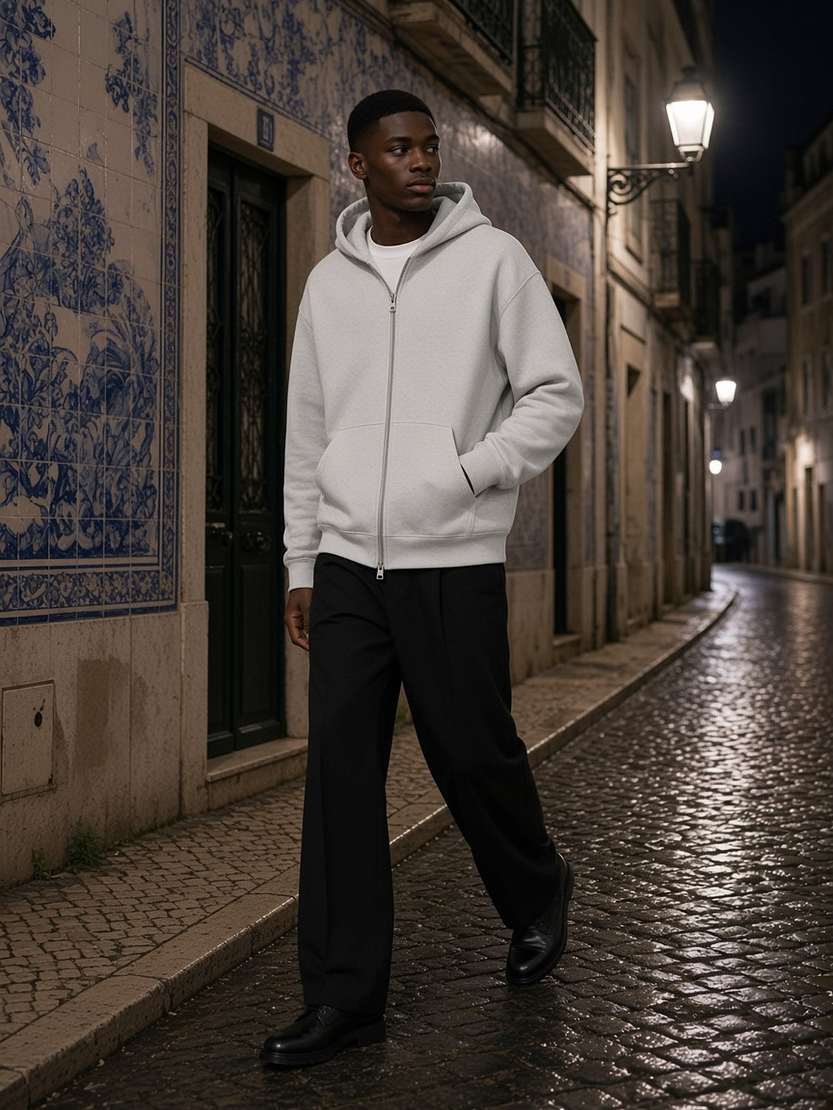 Man in the Men's Heavyweight Full Zip Hoodie in Oat Gray over a white tee, walking a tiled Lisbon lane at night