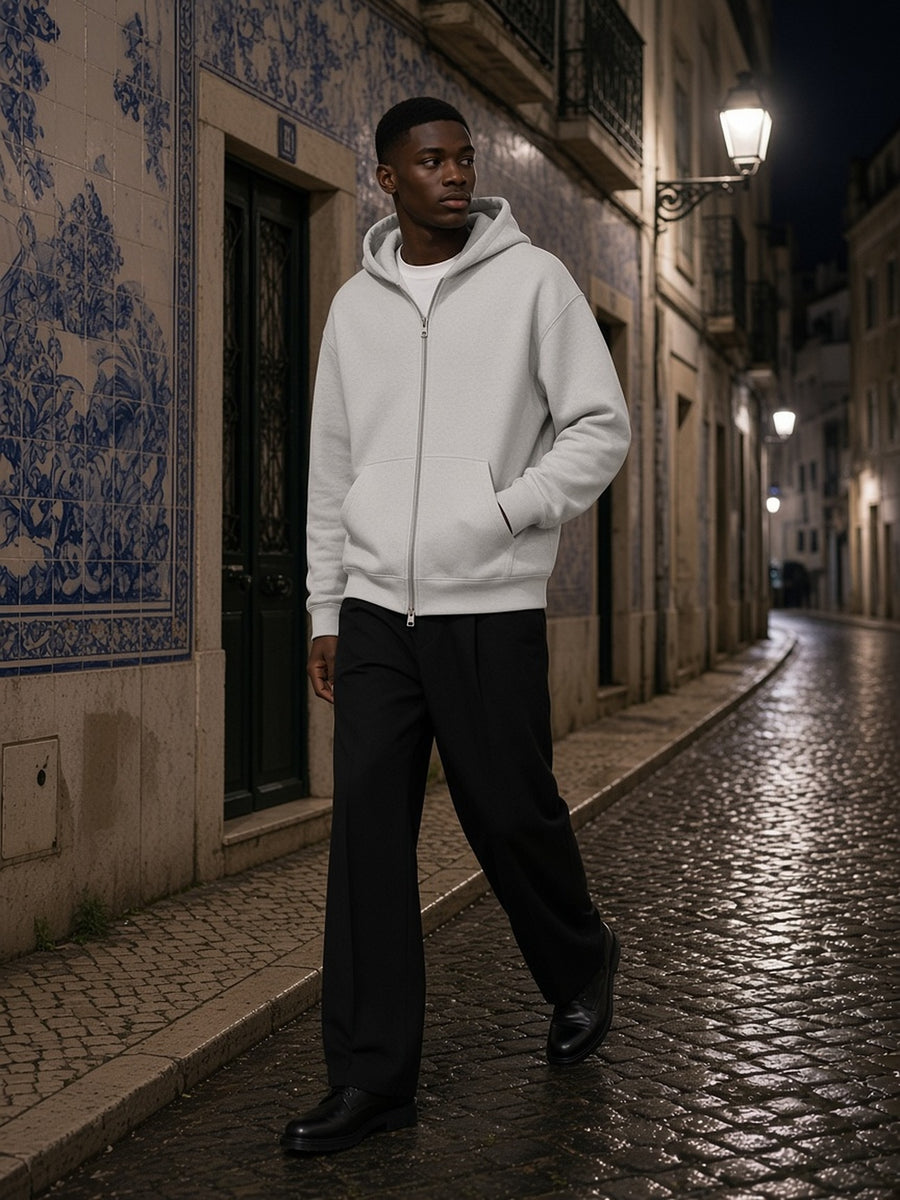 Man in the Men's Heavyweight Full Zip Hoodie in Oat Gray over a white tee, walking a tiled Lisbon lane at night