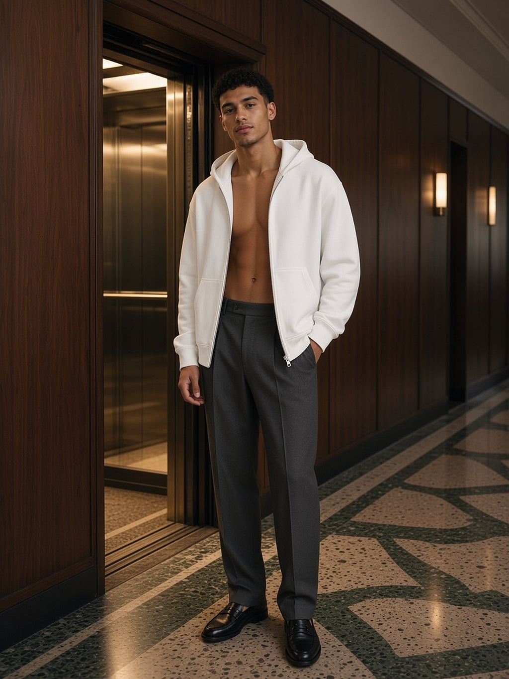 Man in the Men's Heavyweight Full Zip Hoodie in White worn open with charcoal trousers and loafers in a Milan hotel corridor at night