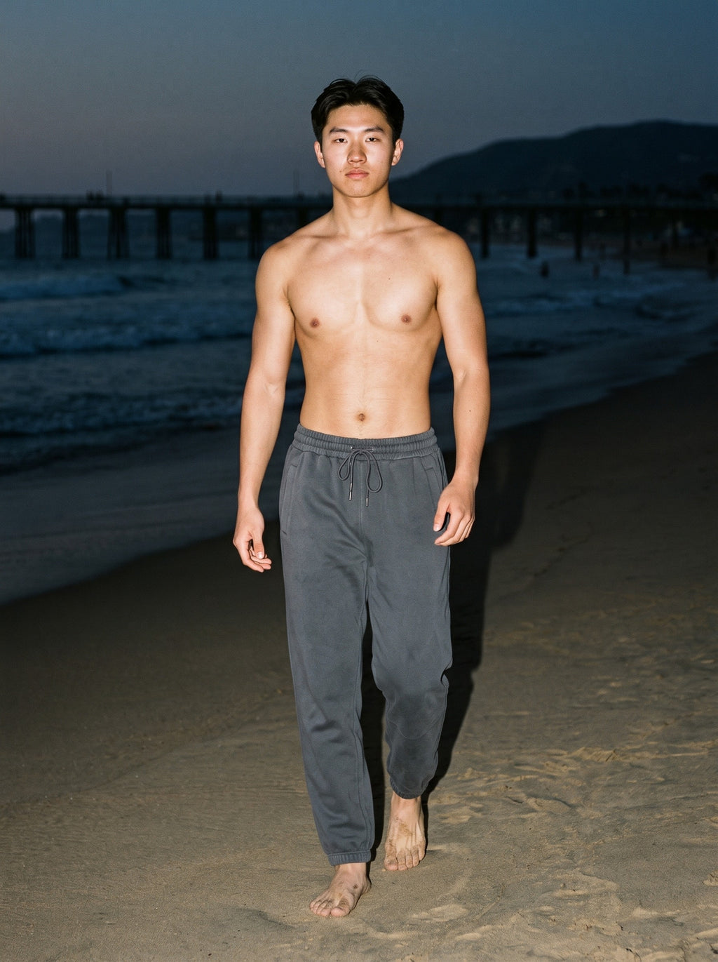 Shirtless man in the Men's Loose Fit Tapered Joggers in Dark Gray, barefoot on the beach after dark, lit by direct flash
