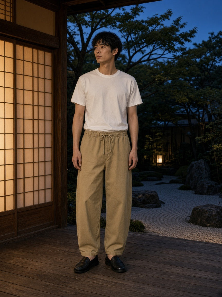 Marcel Afters Mineral Wash Barrel Leg Pants in Khaki, worn by a man, standing in a Japanese courtyard at dusk