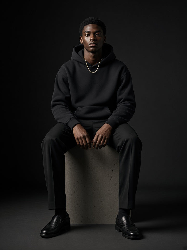 Man in the Oversize Heavyweight Fleece Hoodie in Black Beauty, seated on a concrete plinth in a charcoal studio