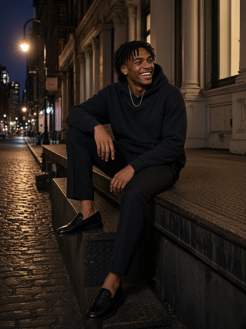 Man in the Oversize Heavyweight Fleece Hoodie in Black Beauty with black trousers and penny loafers, laughing on a SoHo loading dock at night