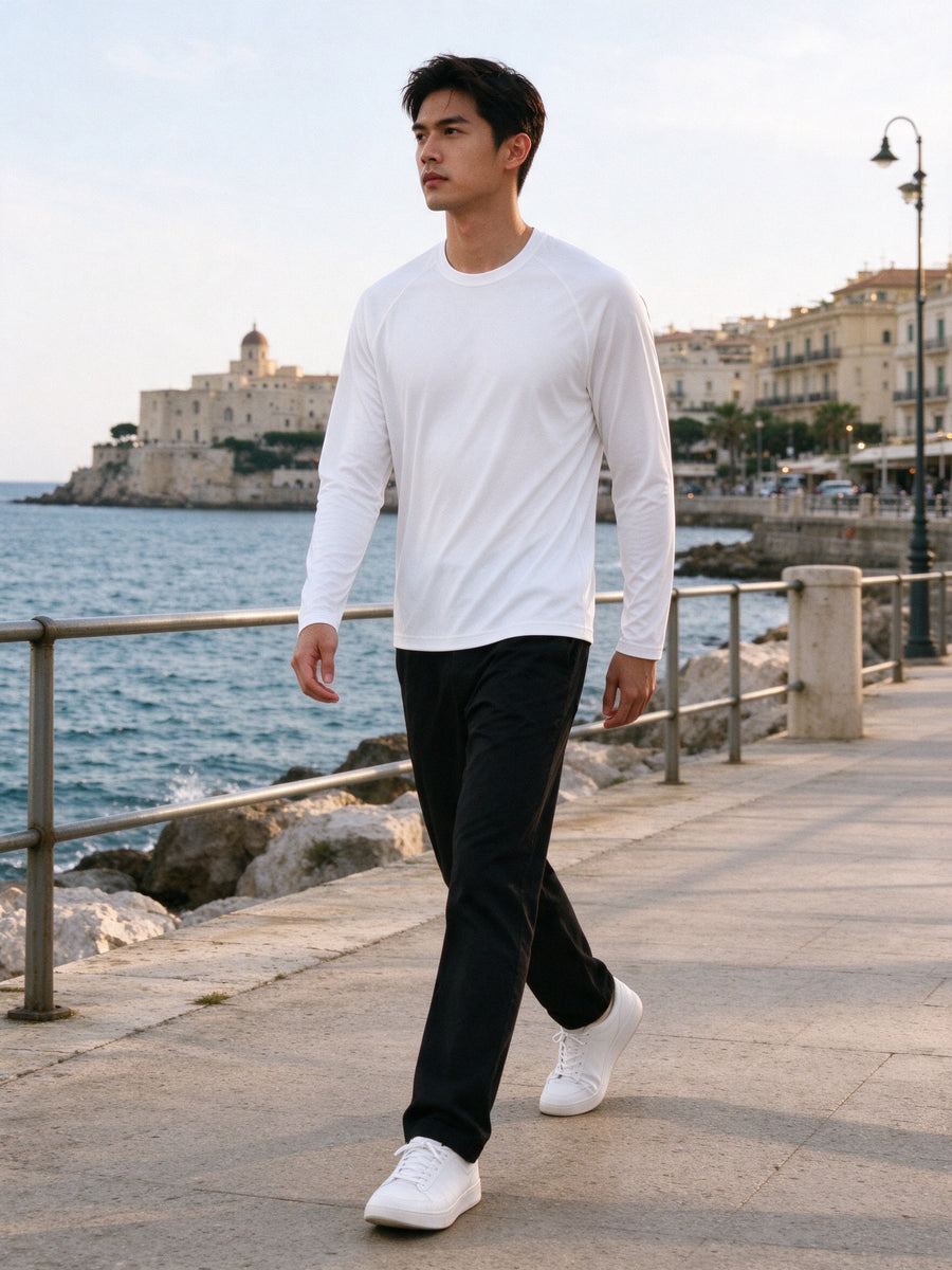 Marcel Afters Performance Long Sleeve T-Shirt in White, worn by a man, striding along a Mediterranean seafront promenade in a white performance long-sleeve tee and black trousers