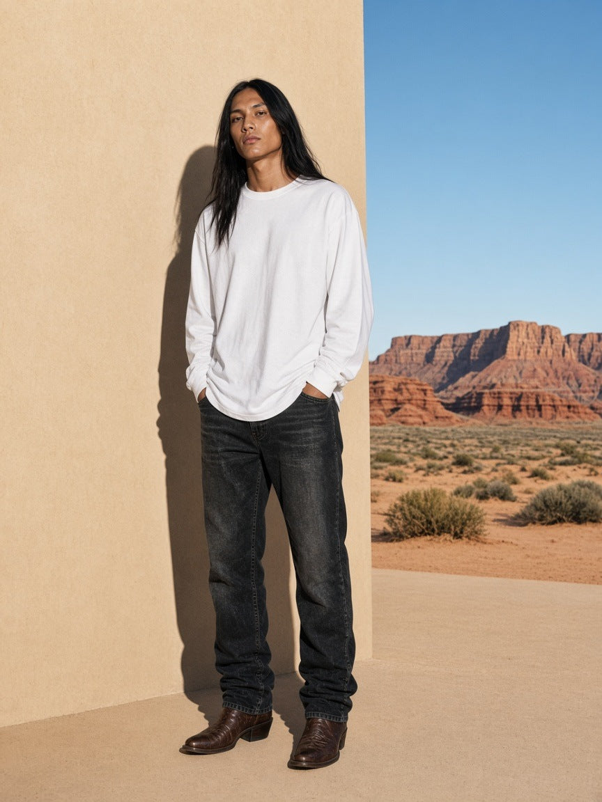 Marcel Afters Raw Hem Long Sleeve T-Shirt in White, worn by a man, standing against a sunlit wall in a white long-sleeve tee and black jeans