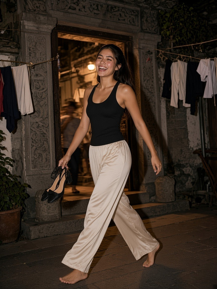 Woman in a black slim fit ribbed racerback tank top and ivory satin trousers, walking barefoot through a lantern-lit courtyard at night