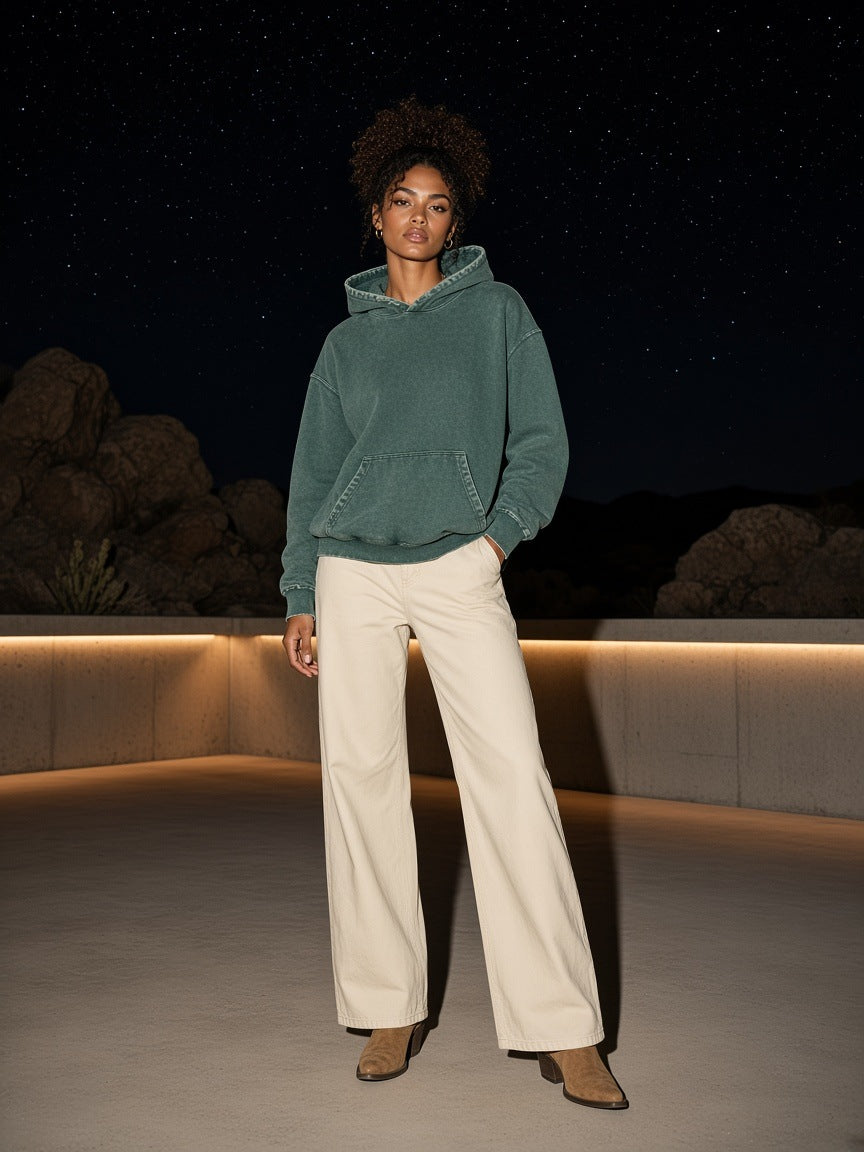 Marcel Afters Snow Wash Hand-Frayed Heavyweight Hoodie in Blackish Green, worn by a woman, standing on a lit terrace at night in wide cream trousers