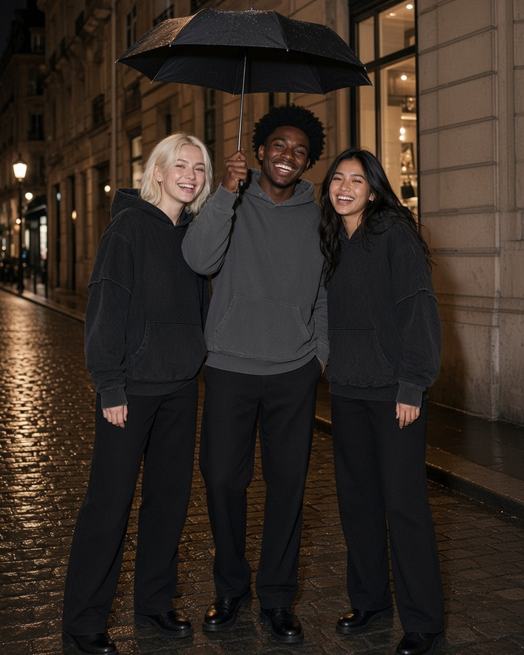 Three people in the Snow Washed Raw Edge Double Layered Hoodie in Black, Dark Gray and Black under one umbrella on a Paris street at night
