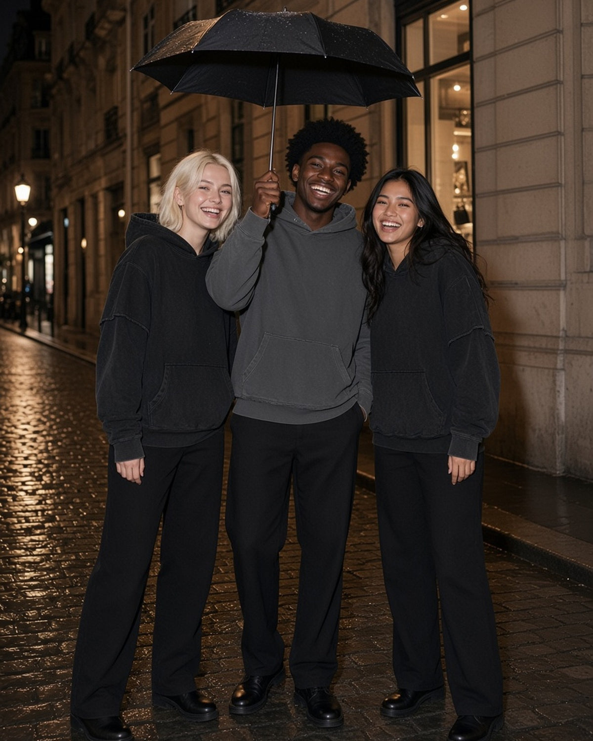 Three people in the Snow Washed Raw Edge Double Layered Hoodie in Black, Dark Gray and Black under one umbrella on a Paris street at night