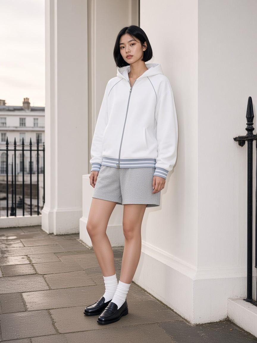 Woman in the Striped Trim Zip-Up Hoodie in White with grey shorts and loafers, on a white stucco portico in London at dawn