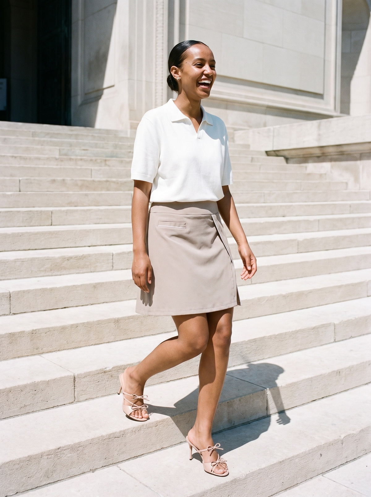 Woman wearing The Bow Tie Sandal in Nude with light coffee high-waisted A-line knee-length skirt with pockets and a white heavyweight oversized polo, flash photograph