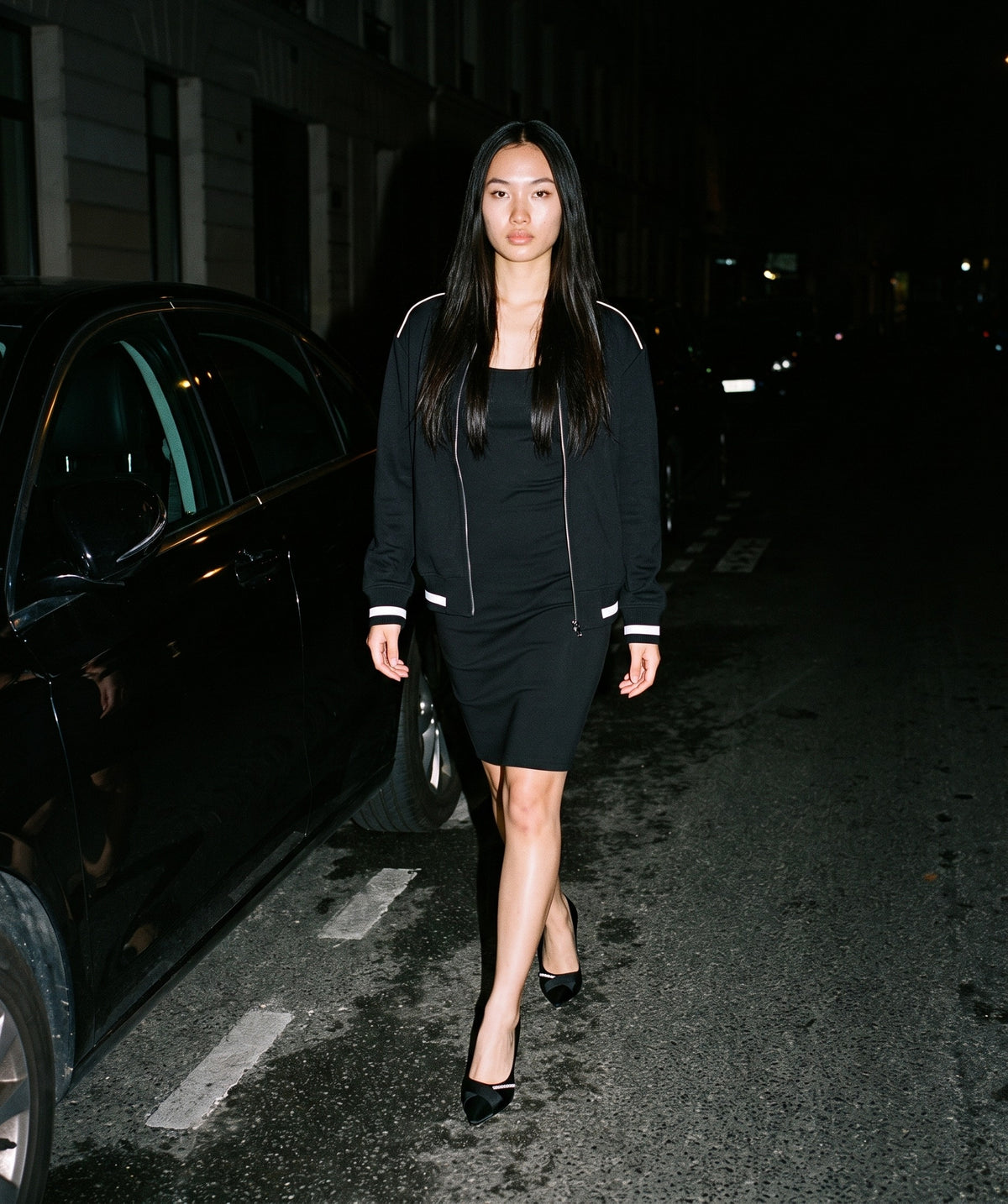 Woman wearing The Cocktail Hour Pump with the Slim Sleeveless Dress in Black, Contrast Piping Zip Bomber Jacket in Black. Past Midnight, Autumn–Winter 2026 campaign