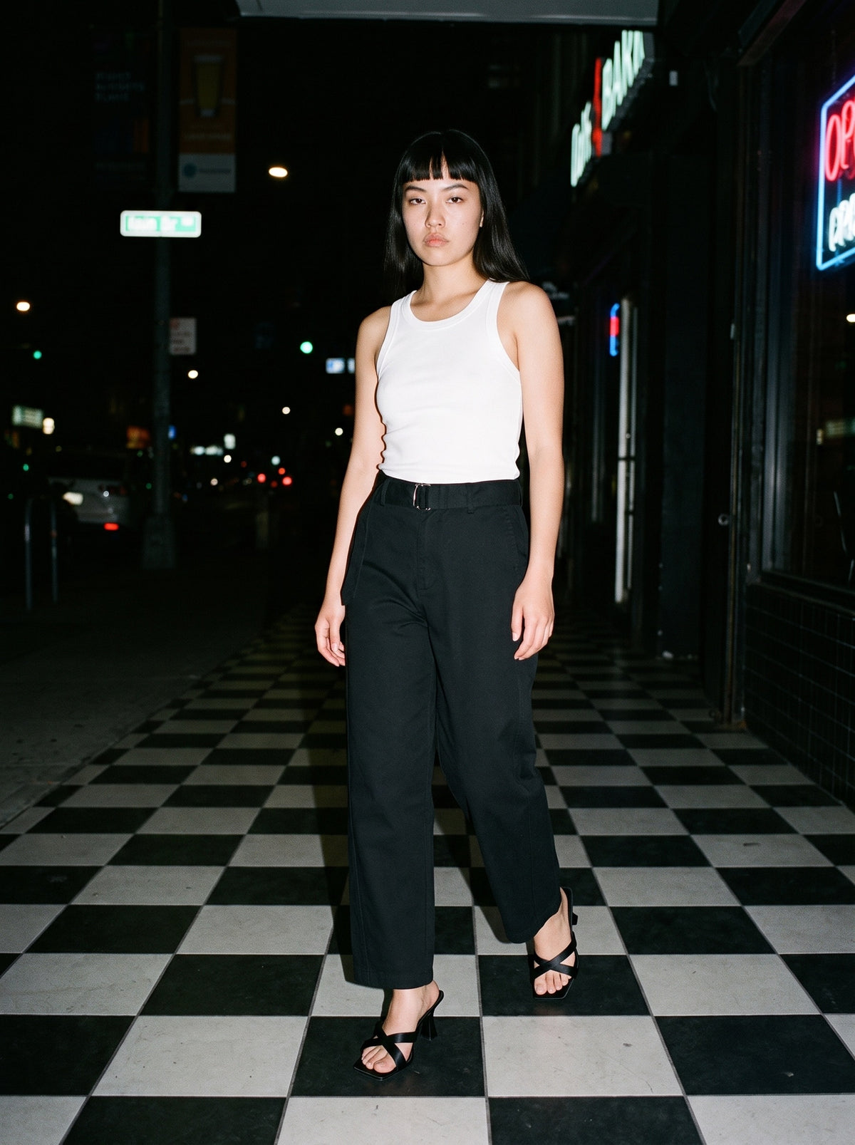 Woman wearing The Crossroads Mule in Black with black belted barrel-leg cotton chino trousers and a white tank top, flash photograph