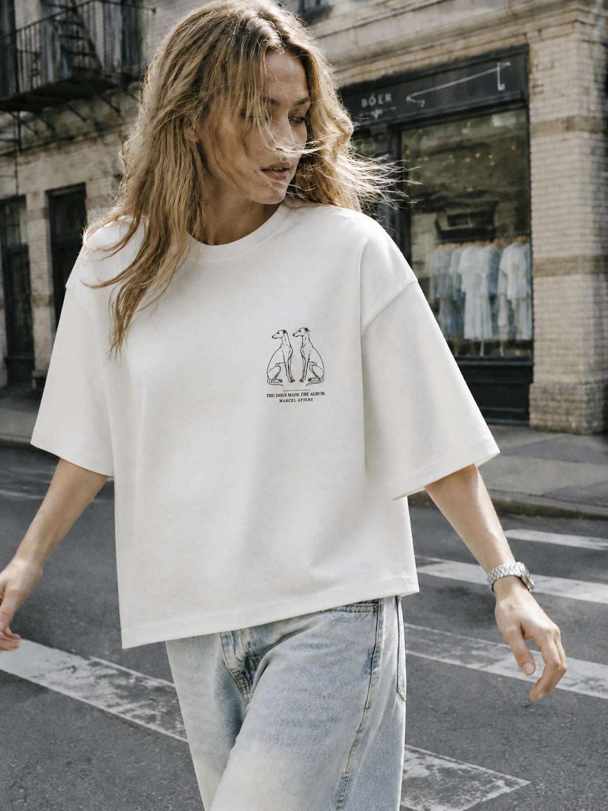 Close-up of a woman in The Dogs Made The Album tee in white, the greyhound drawing on the left chest. Marcel Afters travel wear.