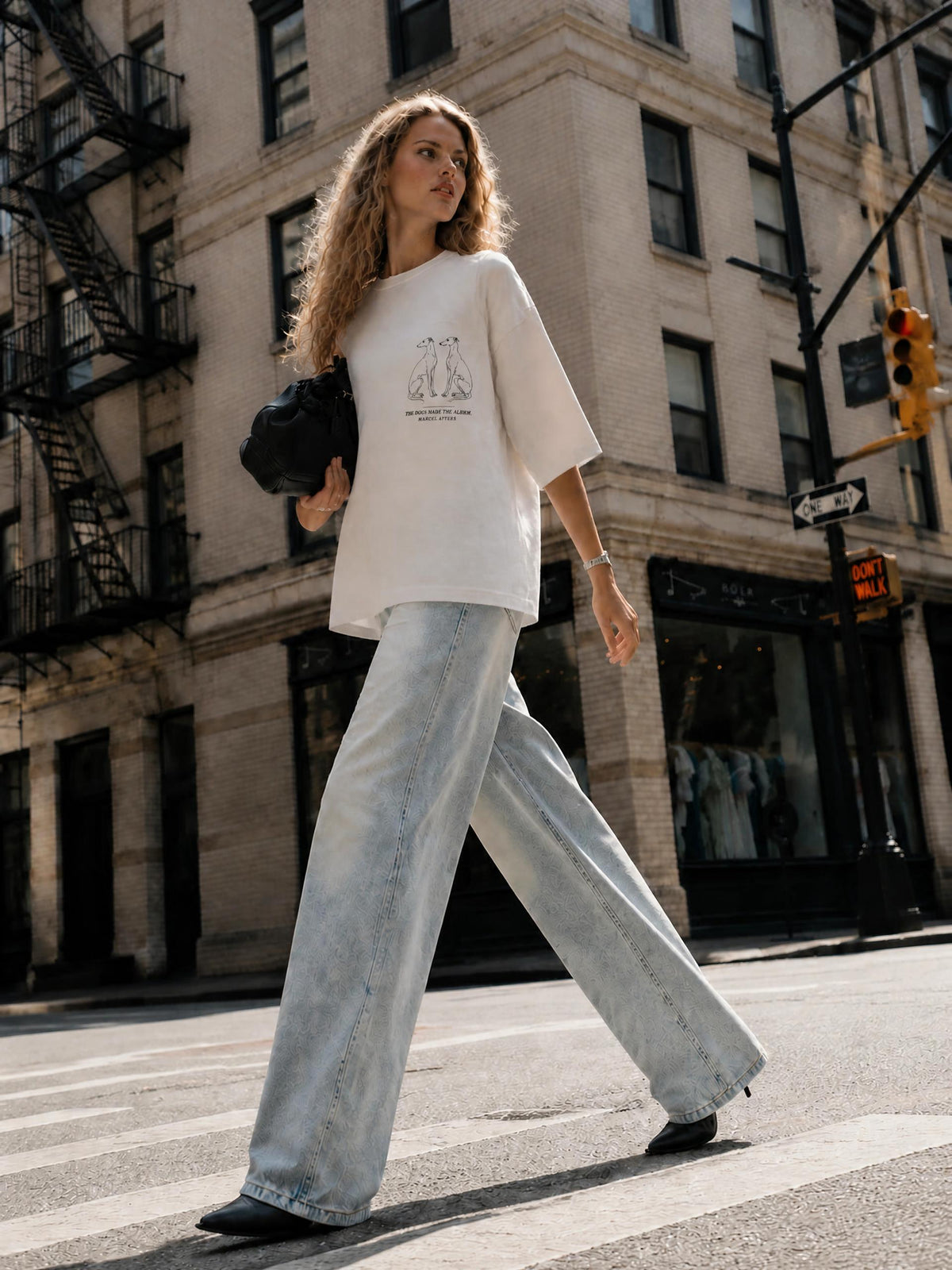 Woman in The Dogs Made The Album heavyweight tee in white with wide-leg jeans, crossing a downtown city street. Marcel Afters.