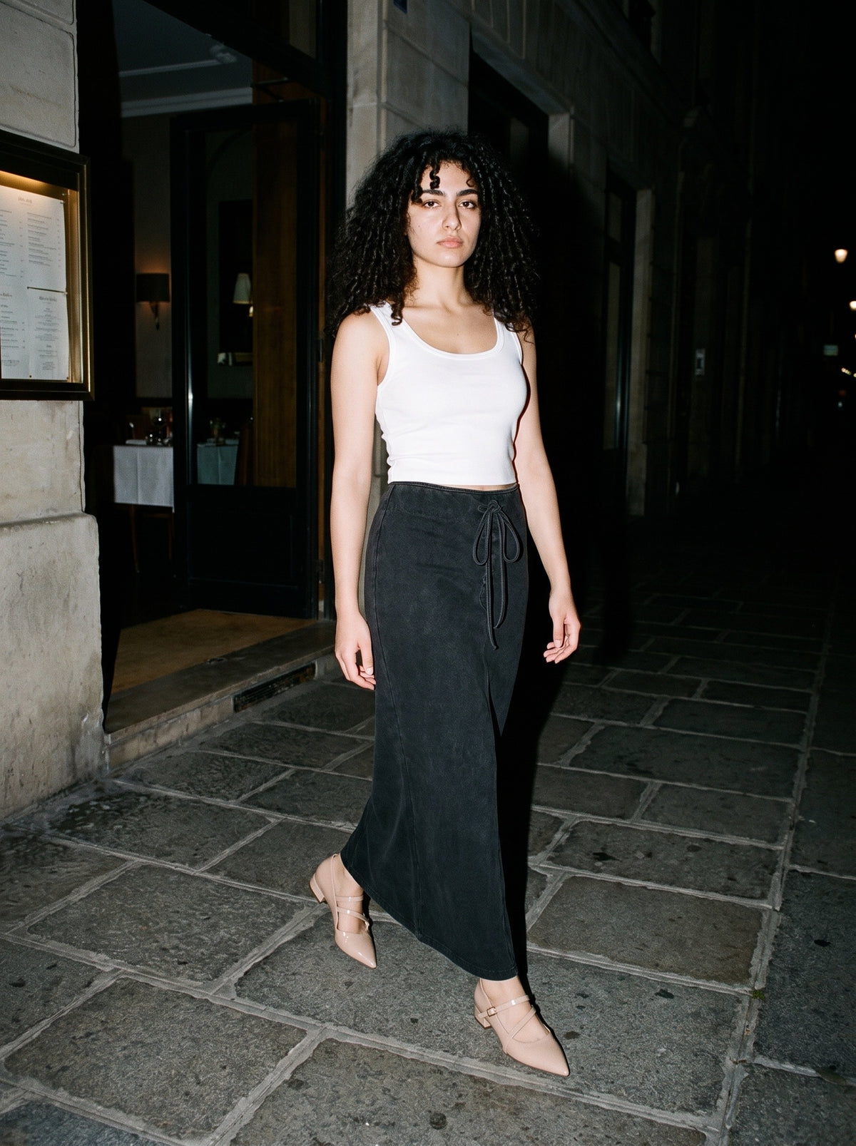 Woman wearing The Gallery Hours Mary Jane in Nude with washed black ankle-length drawstring maxi skirt and a white tank top, flash photograph