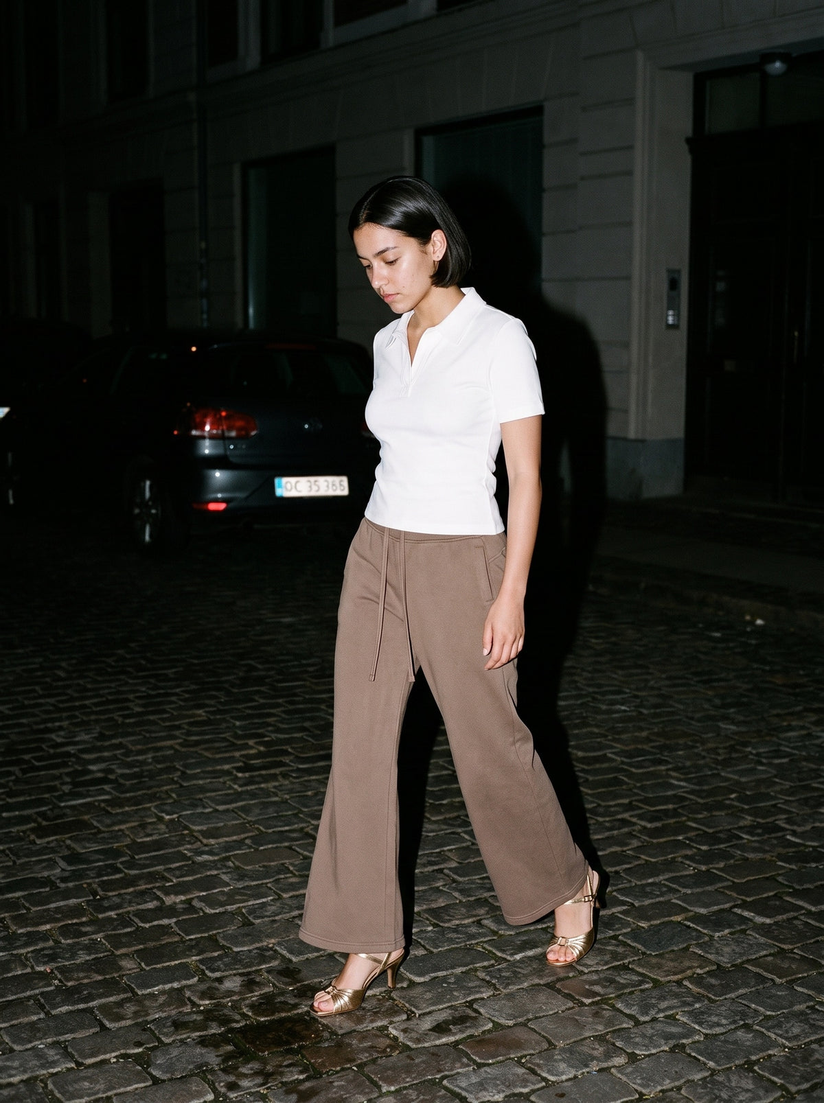 Woman wearing The Golden Hour Mule in Gold with coffee washed flared fleece trousers and a white fitted short-sleeve polo shirt, flash photograph
