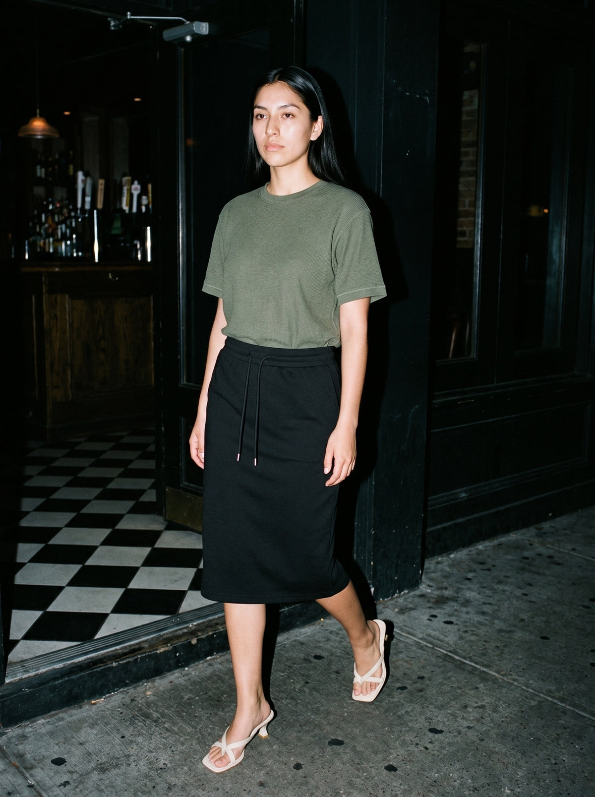 Woman wearing The Hotel Pool Mule in Beige with black midi drawstring-waist jersey skirt and a army green waffle-knit tee, flash photograph
