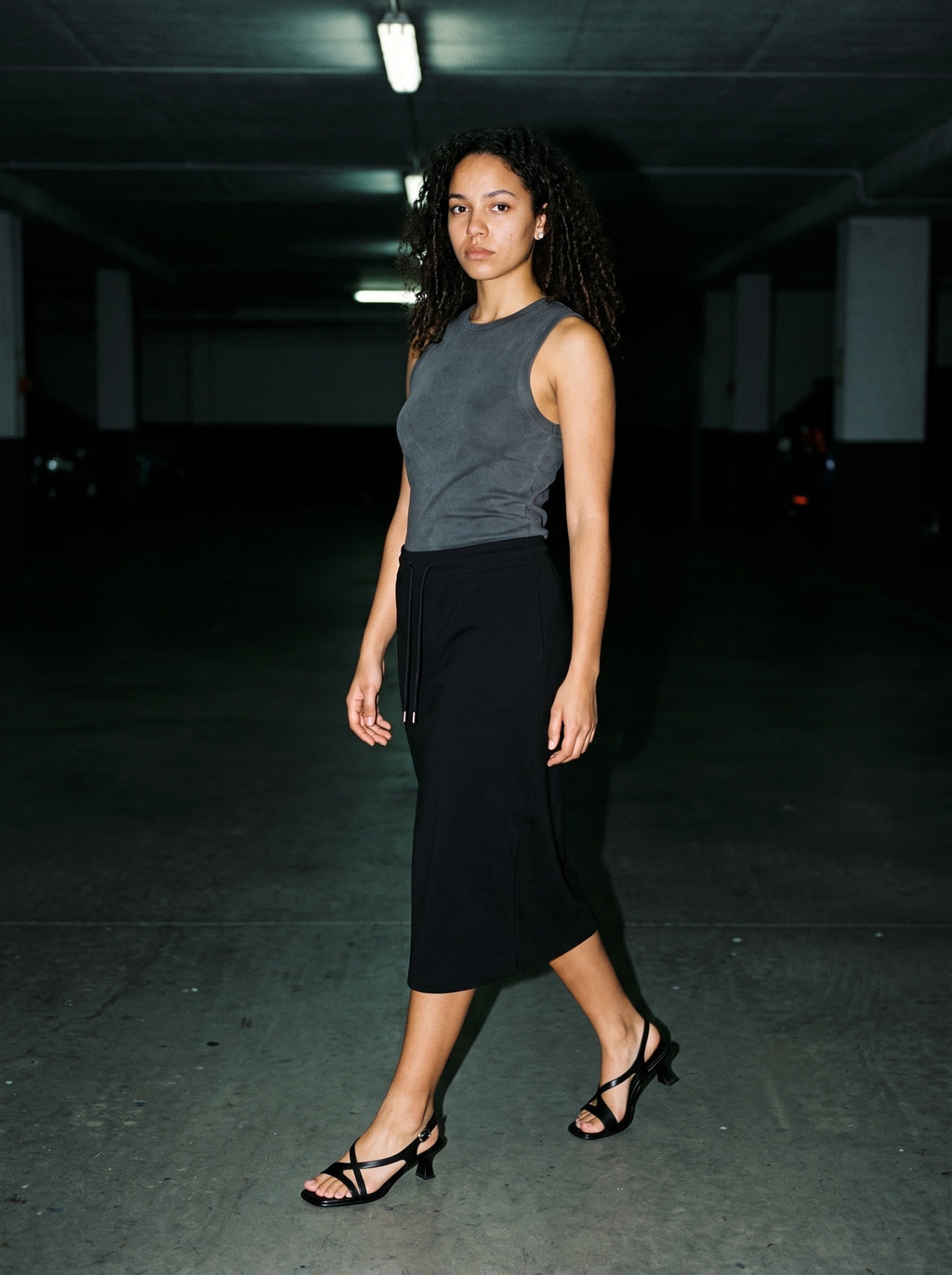 Woman wearing The Late Lunch Sandal in Black with black midi drawstring-waist jersey skirt and a gray tank top, flash photograph