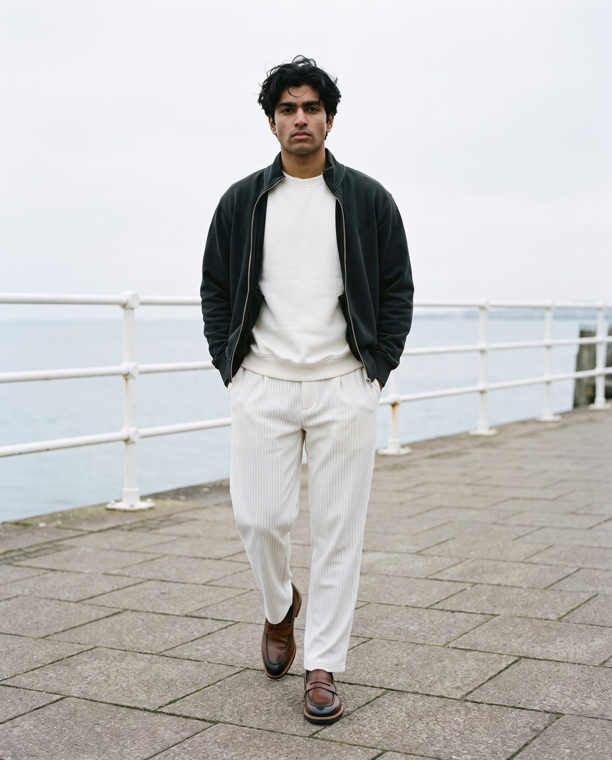 Man wearing The Long Lunch Loafer with the Knitted Round Neck Oversize Sweatshirt in White, Striped Pleated Pants in White, Vintage Wash Fleece Quarter Zip Sweatshirt in Black. The Long Lunch, Autumn–Winter 2026 campaign