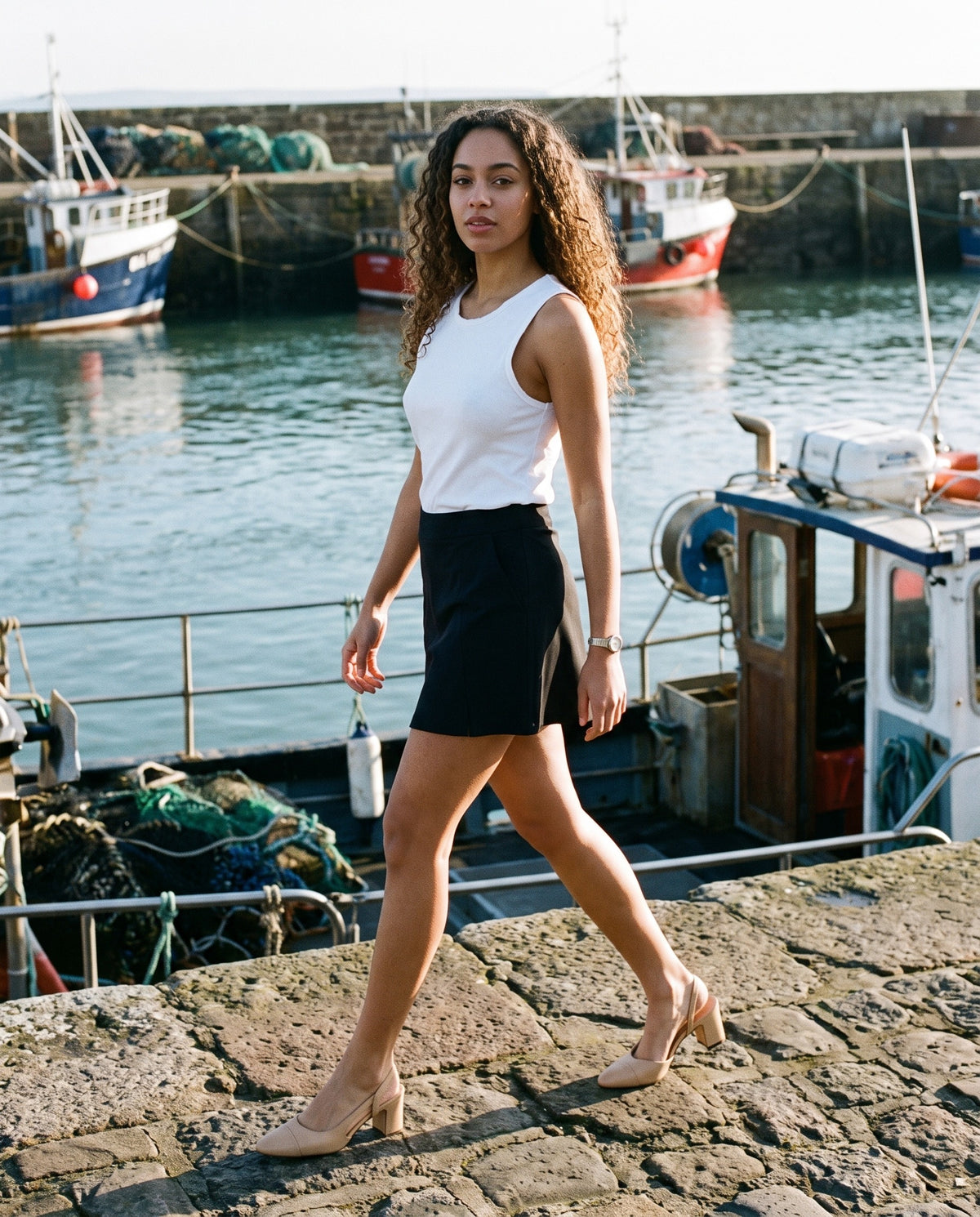 Woman wearing The Long Weekend Slingback with the Raw Edge Cotton Tank Top in White, High-Waist Pocketed A-Line Skirt in Black. Travel Light, Autumn–Winter 2026 campaign