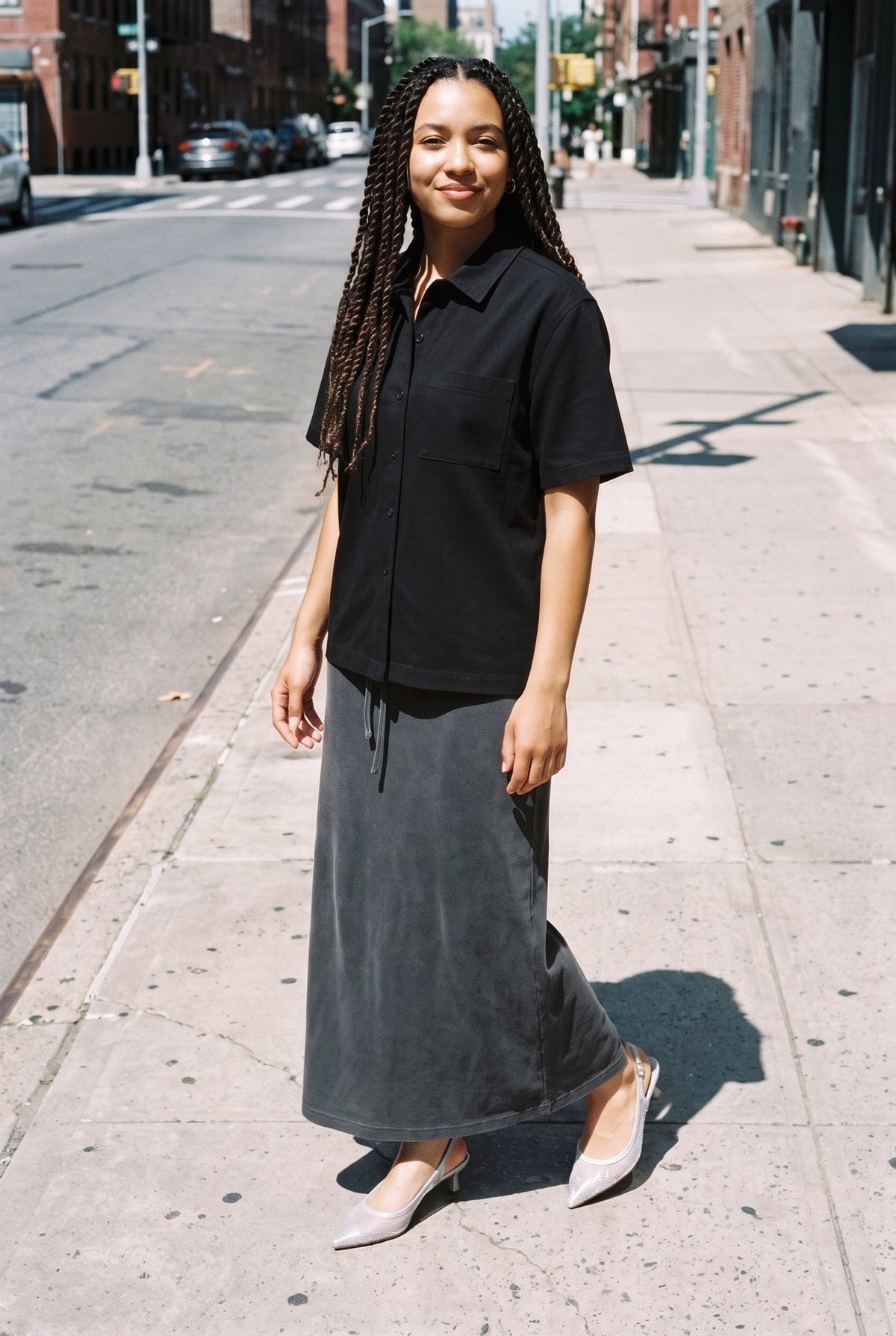 Woman with long braids wearing The Moonlight Slingback in silver with a black short-sleeve shirt and a washed charcoal maxi skirt, full length on a sunlit city street