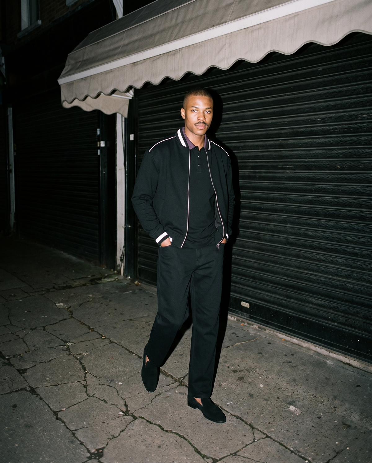 Man wearing The Night Porter Slipper with the Contrast Collar Raglan Polo Shirt in Black, Heavyweight Straight-Leg Trousers in Black, Contrast Piping Zip Bomber Jacket in Black. After Dark, Autumn–Winter 2026 campaign