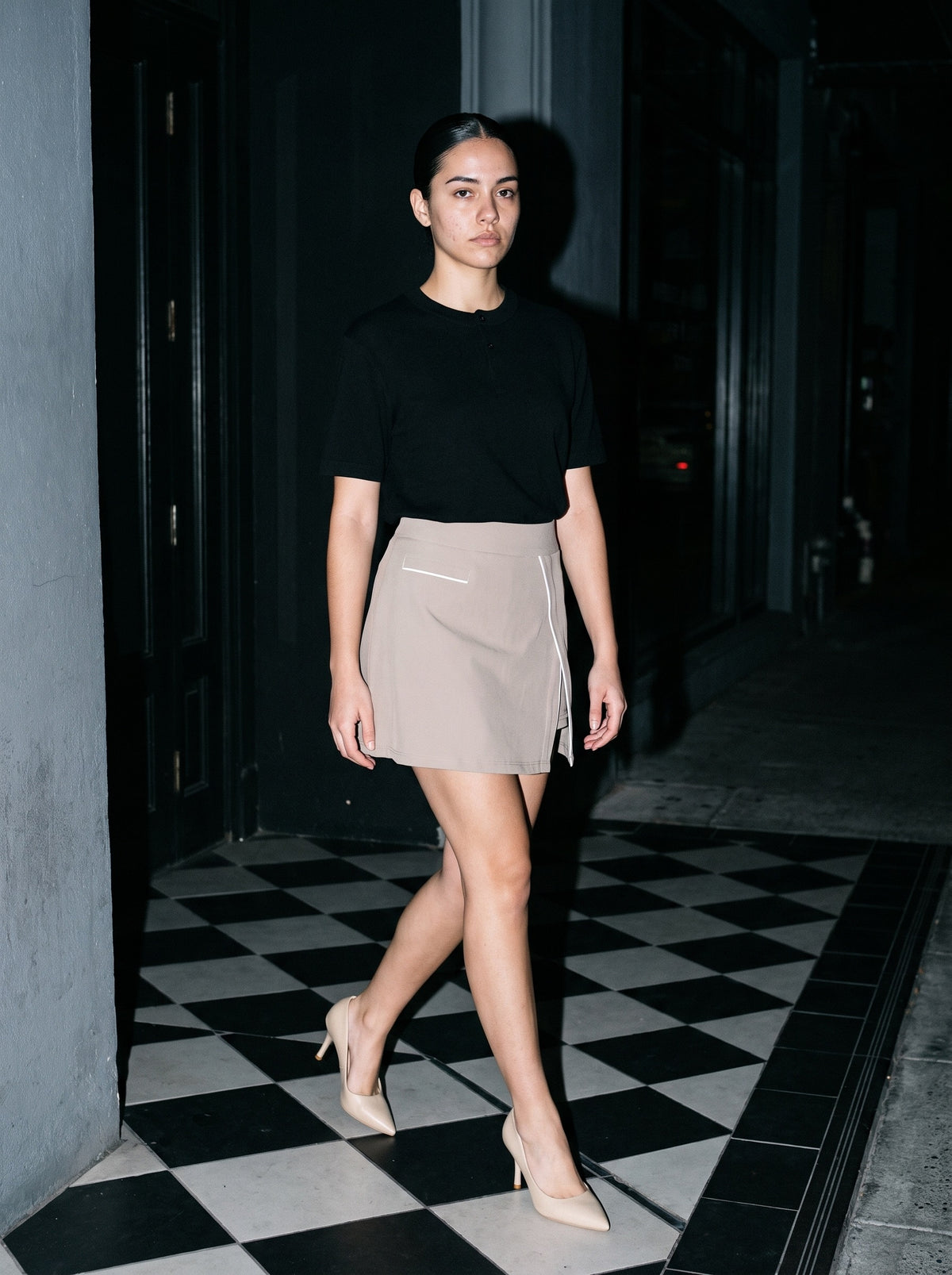 Woman wearing The Reservation Pump in Nude with light coffee high-waisted A-line knee-length skirt with pockets and a black short-sleeve knit henley tee, flash photograph