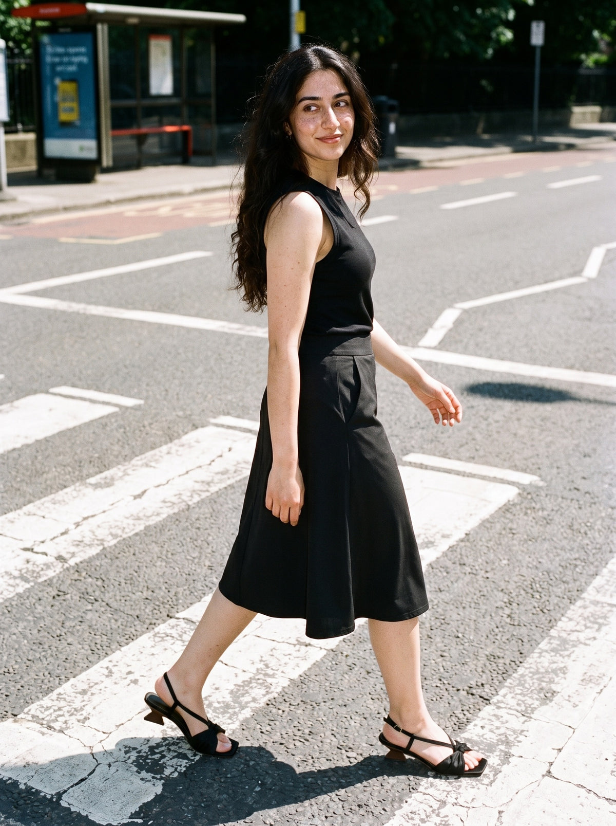 Woman wearing The Rooftop Sandal in Black with black high-waisted A-line knee-length skirt with pockets and a black tank top, flash photograph