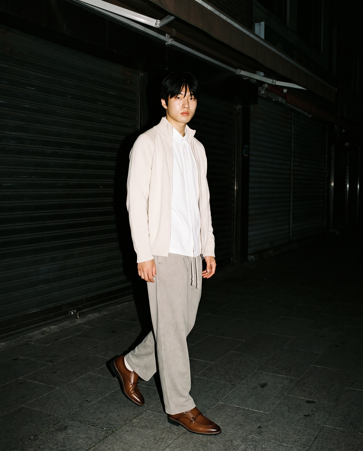 Man in The Second Bottle Monk in Brown with oat trousers and a cream knit jacket, walking a shuttered street at night