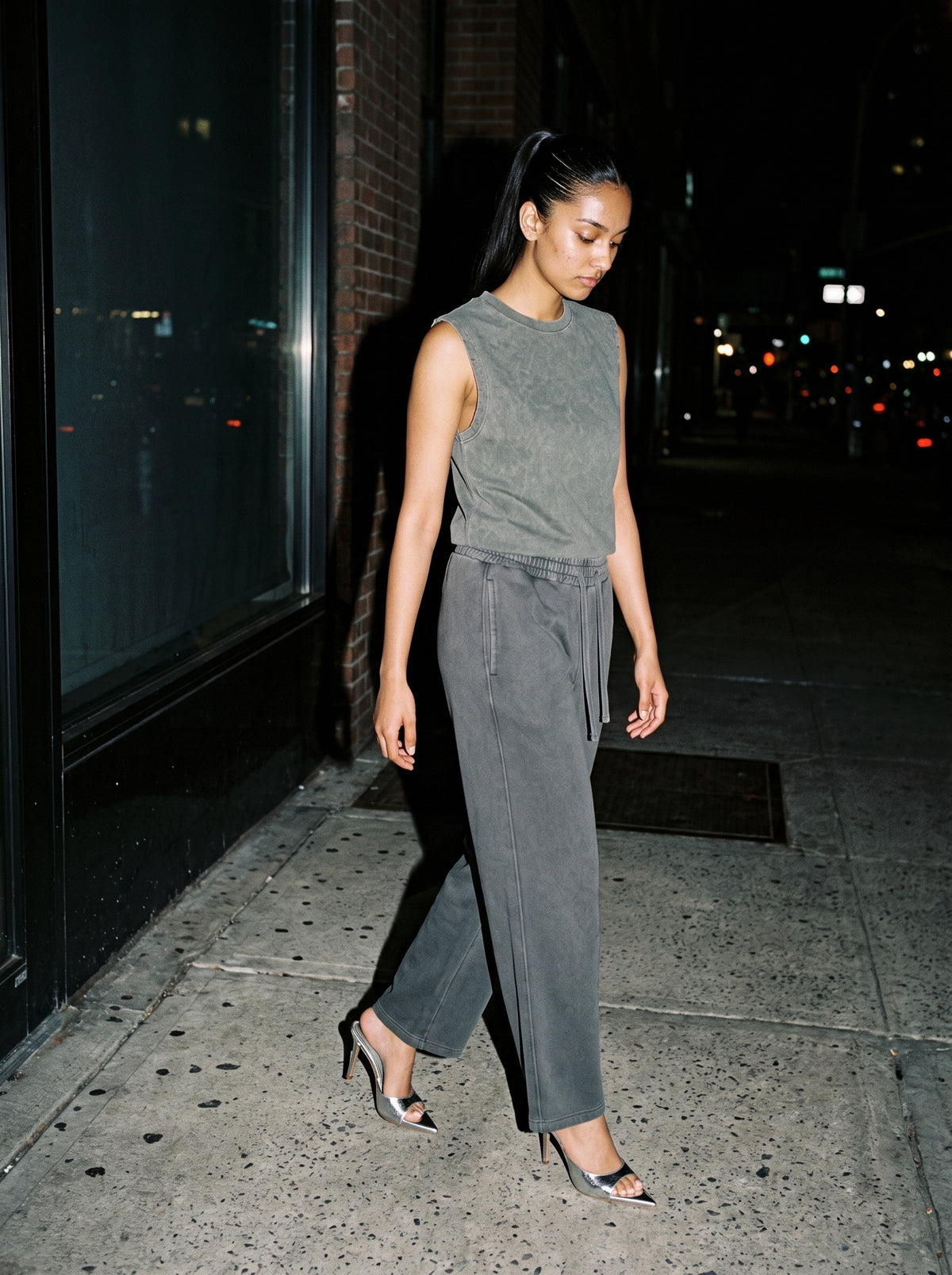 Woman wearing The Silver Screen Mule in Silver with dark gray vintage-washed straight-leg wide-waist trousers and a gray green tank top, flash photograph