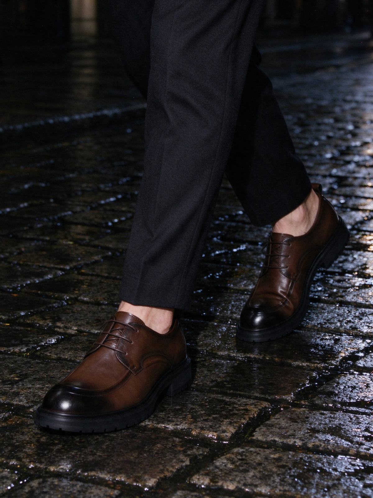 The Wet Pavement Derby in brown on foot, wet cobblestone street at night