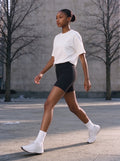 Marcel Afters Heavyweight Boxy T-Shirt in White, worn by a woman, walking a sunlit city square in a white heavyweight boxy tee and black cycling shorts