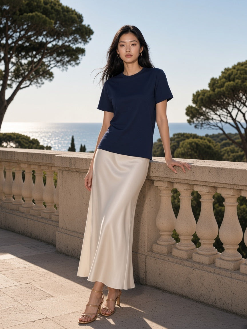 Marcel Afters Solid Color Regular Fit T-Shirt in Navy Blue, worn by a woman, leaning on a stone balustrade by the sea in a satin skirt