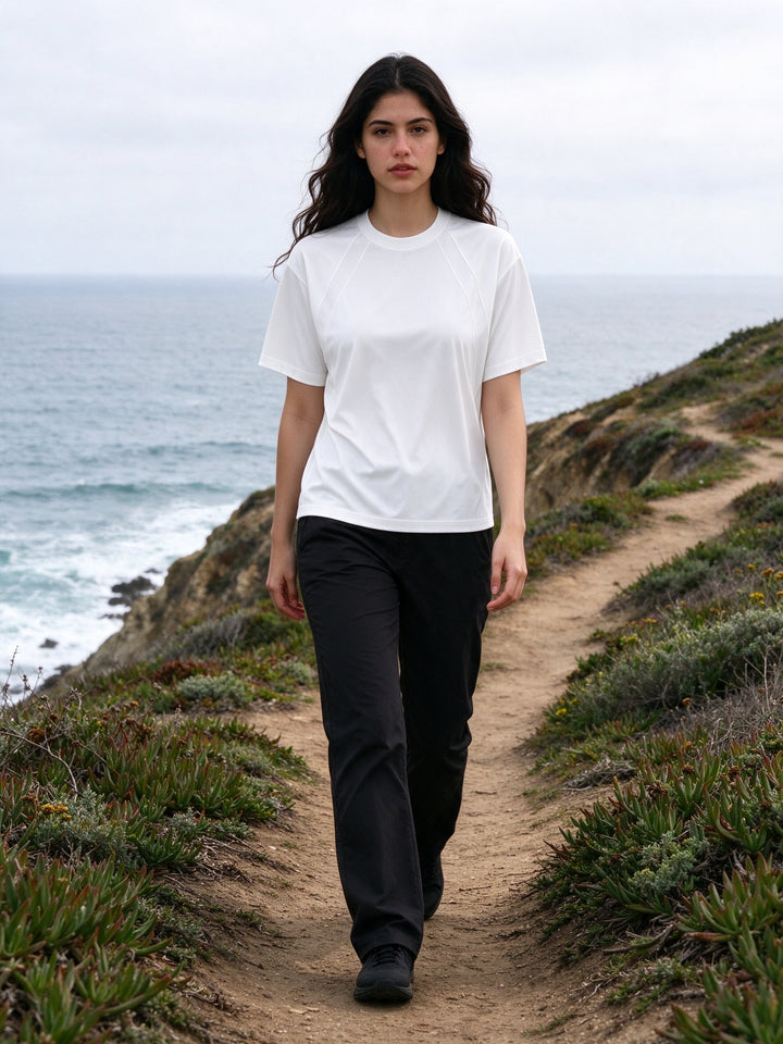 Marcel Afters Tonal Paneled Performance T-Shirt in White, worn by a woman, walking a clifftop coastal path above the sea in a white tonal-paneled performance tee and black trousers