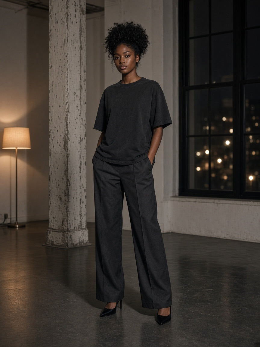 Marcel Afters Vintage Wash Drop Shoulder T-shirt in Black, worn by a woman, standing in a loft with city lights behind, in wide trousers