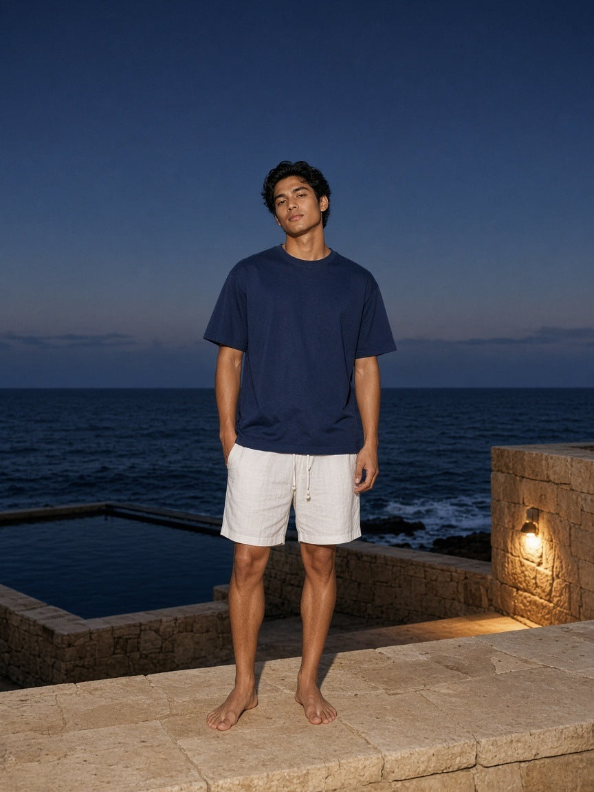 Marcel Afters Vintage Wash Drop Shoulder T-shirt in Navy Blue, worn by a man, standing barefoot on a stone terrace above the sea at dusk