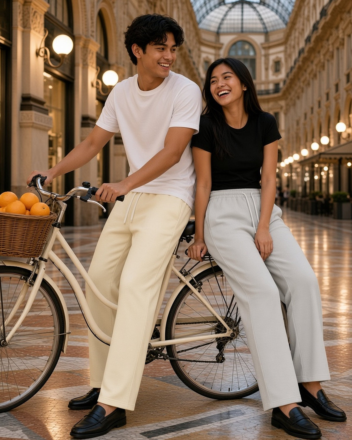 Two people in the Waffle Barrel Leg Pants in Oat Gray and Mild Apricot with a vintage bicycle in a Milan arcade at dusk