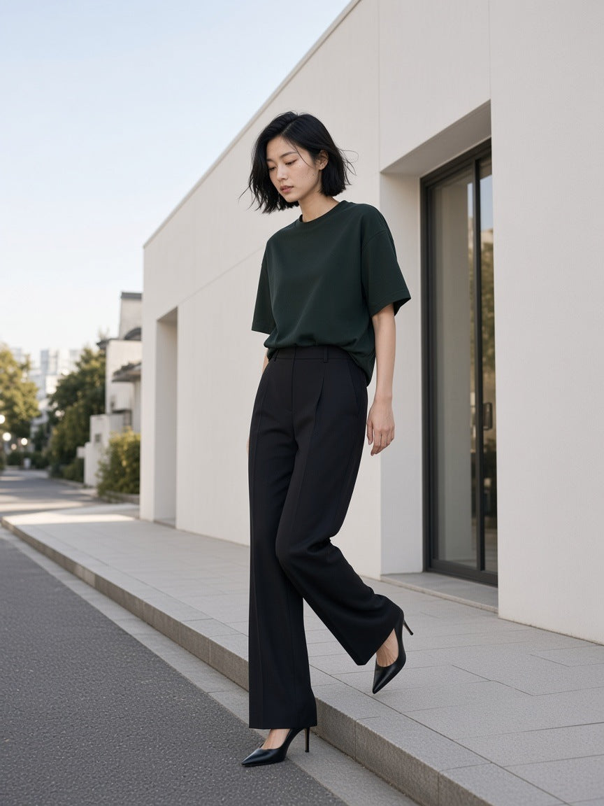 Marcel Afters Women's Heavyweight Boxy T-Shirt in Blackish Green, worn by a woman, stepping along a white wall in tailored black trousers