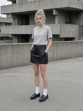 Woman in the Women's Heavyweight Earth Tone T-Shirt in Oat Gray with a black skirt, brutalist concrete courtyard