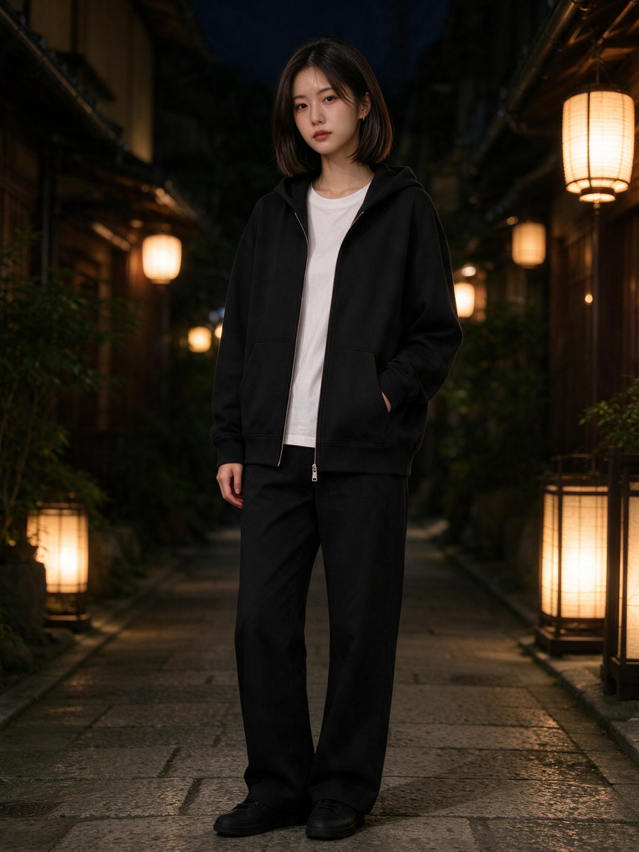 Marcel Afters Women's Heavyweight Full Zip Hoodie in Black, worn by a woman, standing in a lantern-lit Kyoto alley at night in a black full-zip hoodie over a white tee and black trousers