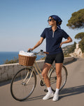 Woman in the Women's High Waisted Yoga Biker Shorts in Black with a navy quarter-zip polo and a silk head scarf, beside a vintage bicycle on a Riviera coast road