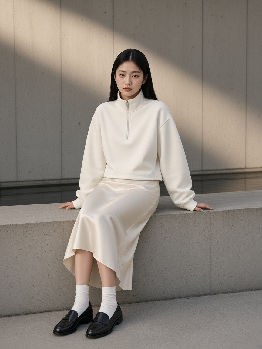 Woman in the Women's Mock-Neck Quarter-Zip Sweatshirt in White with an ivory silk skirt, seated in a raw concrete courtyard in Tokyo