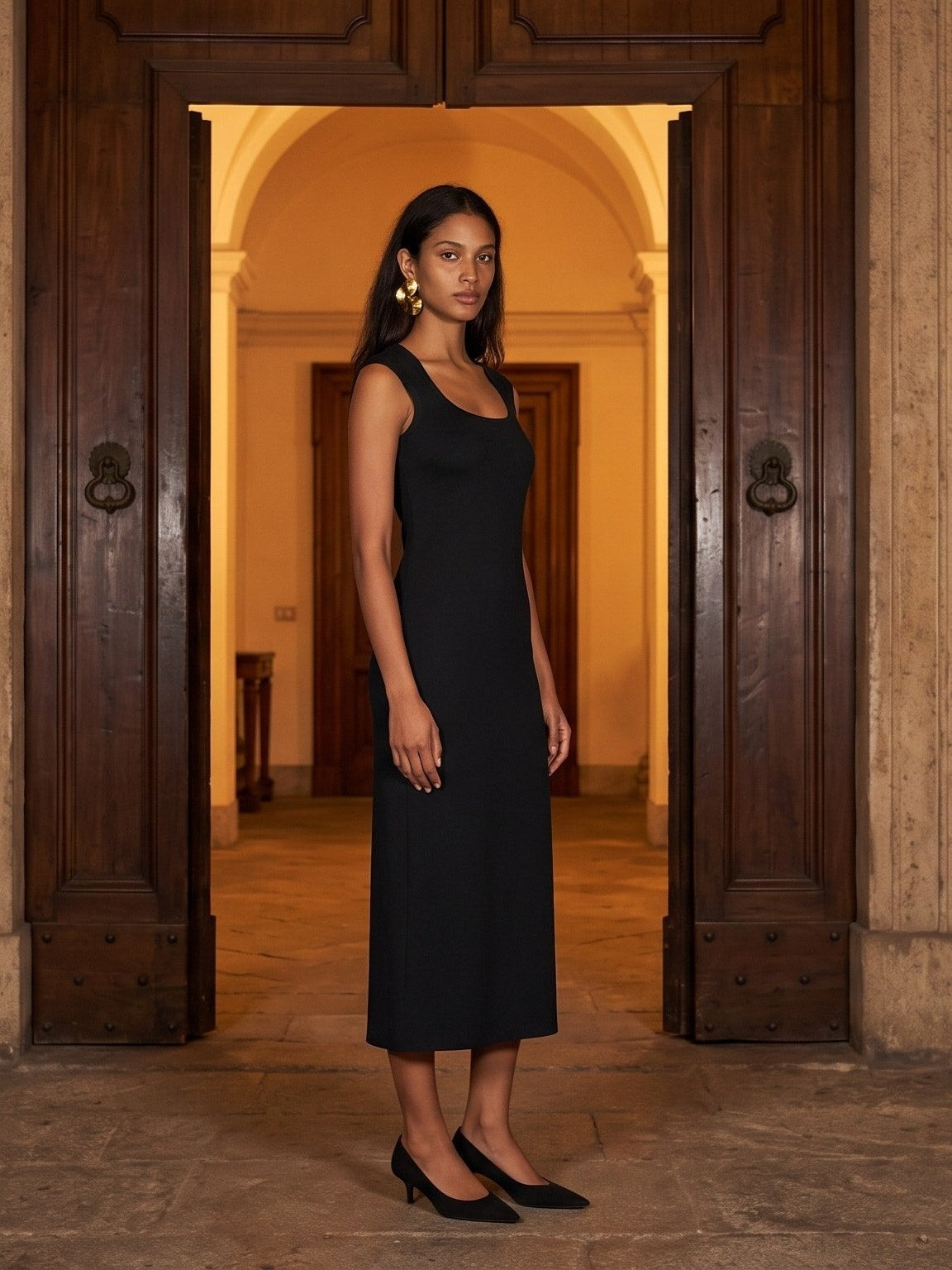 Woman in the Women's Slim Sleeveless Dress in Black, scoop neck and mid-calf length, with black pointed heels, standing in an arched doorway between tall wooden doors in a warmly lit stone hall