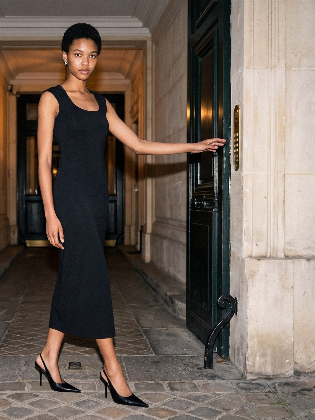 Woman in the Women's Slim Sleeveless Dress in Black, scoop neck and mid-calf length, with black pointed heels, walking through a stone courtyard passage between open black doors