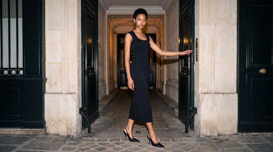 Woman in the Marcel Afters Women's Slim Sleeveless Dress in Black, scoop neck and mid-calf length, with black pointed heels, walking through a stone courtyard passage between open black doors