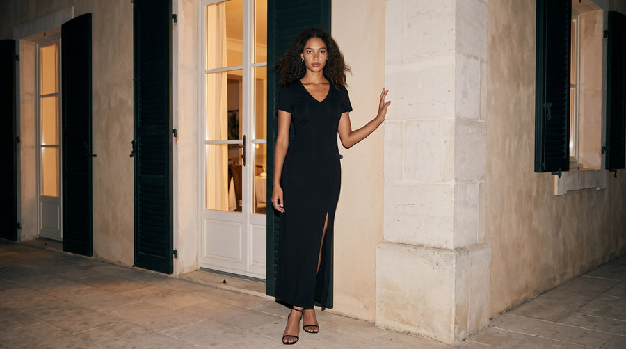 Woman in the Marcel Afters Women's V-Neck Short Sleeve Maxi Dress in Black with a front slit and black sandals, standing by tall French windows and stone walls at night
