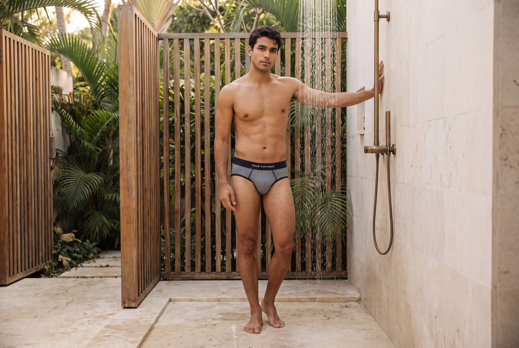 Man wearing the True Notions Tartan Brief — Grey, standing by a slatted outdoor shower, full-length editorial photograph