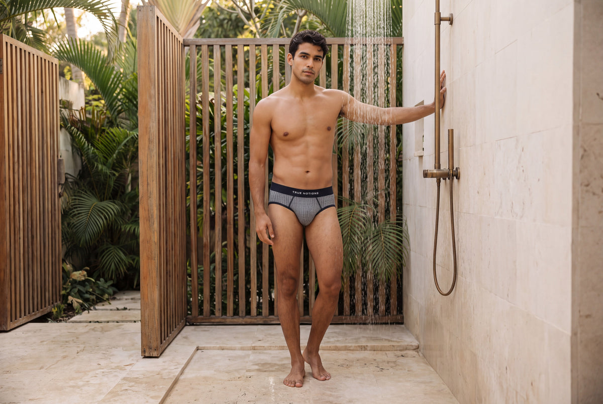 Man wearing the True Notions Tartan Brief — Grey, standing by a slatted outdoor shower, full-length editorial photograph