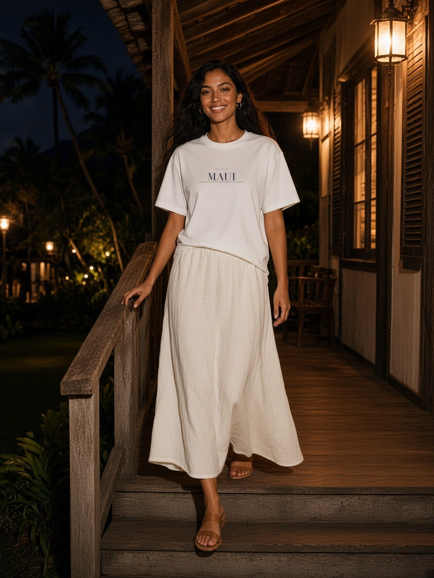Woman in the Maui Heavyweight Drop Shoulder T-Shirt in White with the navy Maui city plate, on a torchlit beach path under the palms in Maui at night